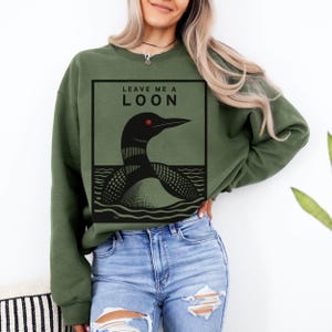 May include: A green sweatshirt with a black and white illustration of a loon on the front. The text "Leave Me A Loon" is printed above the loon.