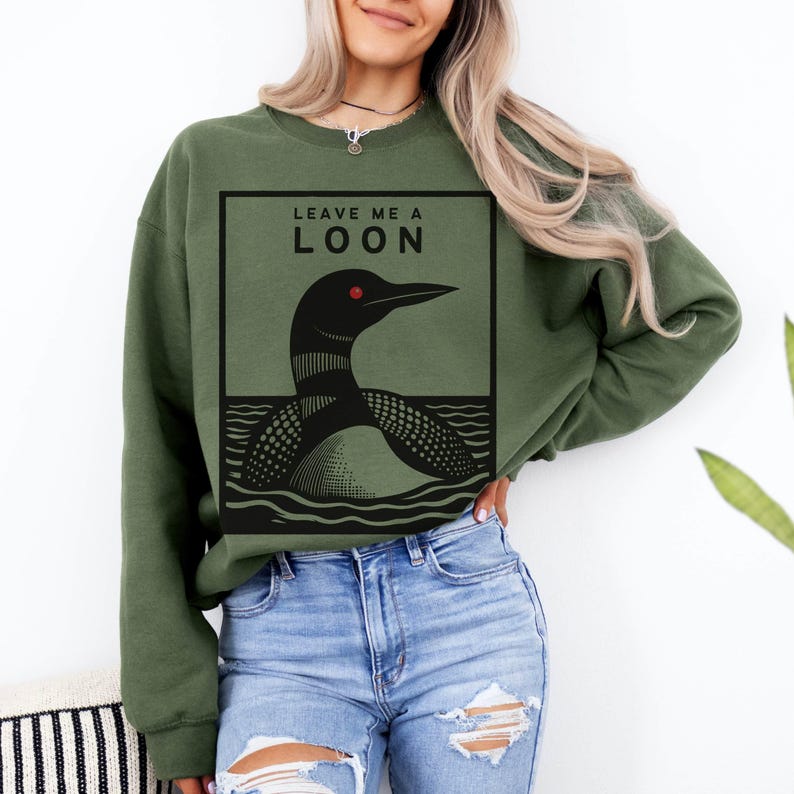 May include: A green sweatshirt with a black and white illustration of a loon on the front. The text "Leave Me A Loon" is printed above the loon.