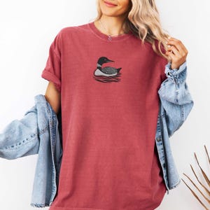 May include: A red short-sleeved t-shirt with a black and white loon graphic on the front. The loon is swimming in water.