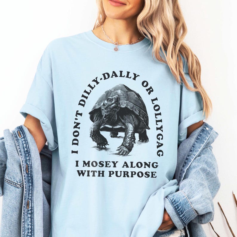 May include: Light blue t-shirt with a black and white graphic of a tortoise. The graphic says "I don't dilly-dally or lollygag I mosey along with purpose".