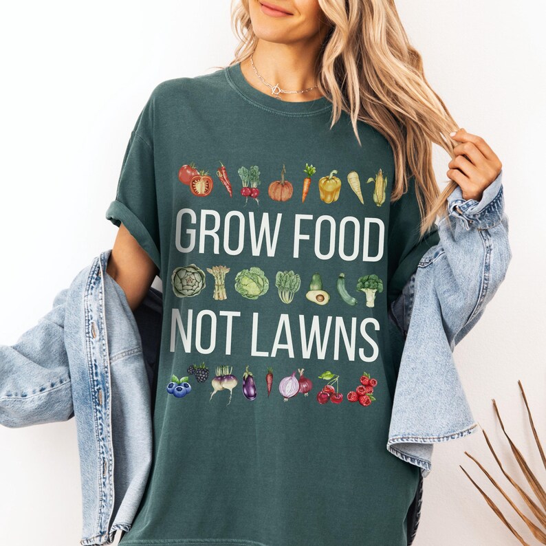 May include: Dark green t-shirt with the text "GROW FOOD NOT LAWNS" in white. The shirt features watercolor illustrations of various vegetables and fruits, including tomatoes, peppers, and berries. The shirt is a comfortable, casual style.
