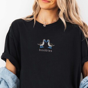 May include: Black t-shirt featuring an embroidered design of two blue-footed boobies facing each other. The word "boobies" is embroidered below the birds. The shirt is a casual, unisex style.