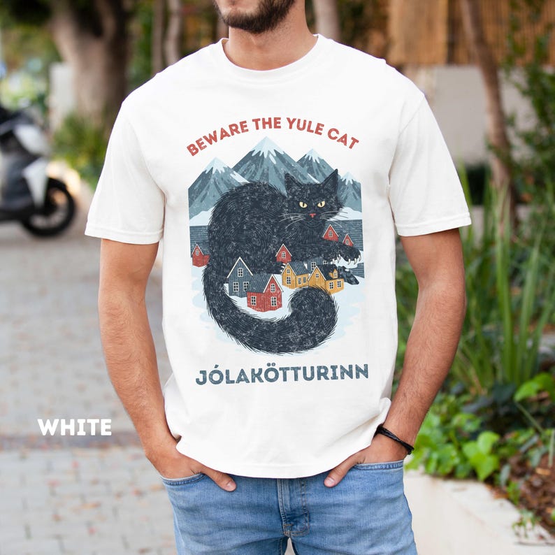 May include: White t-shirt with a graphic of a black cat, mountains, and colorful houses. Text on the shirt reads "BEWARE THE YULE CAT" and "J&Oacute;LAK&Ouml;TTURINN". The word "WHITE" is printed at the bottom left.
