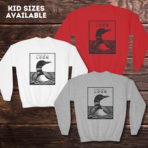 May include: Three sweatshirts in red, white, and gray with a black and white illustration of a loon on each. The loon has red eyes and the text "Leave me a loon" is printed above the loon.