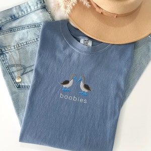 May include: A blue Comfort Colors t-shirt with an embroidered design of two blue-footed boobies and the word "boobies" in white. The shirt is laid on top of a pair of light-wash jeans, a tan hat, and a gold necklace.