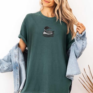 May include: A dark green t-shirt with a black and white loon graphic. The loon is swimming in water with ripples.