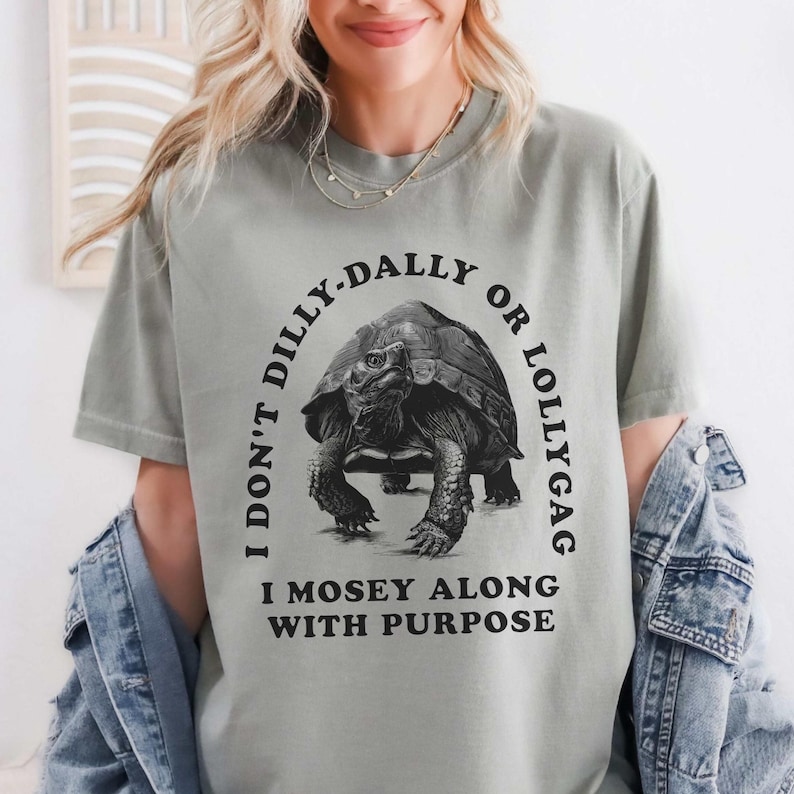 May include: A gray t-shirt with black text that reads 'I don't dilly-dally or lollygag I mosey along with purpose.' A black and white illustration of a tortoise is in the center of the text.
