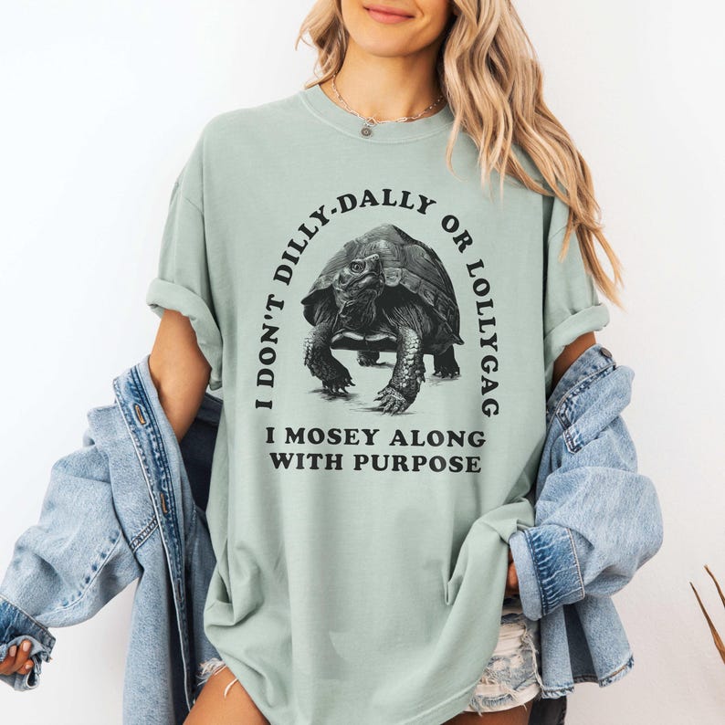 May include: A light green t-shirt with a black graphic of a tortoise and the text "I don't dilly-dally or lollygag I mosey along with purpose".