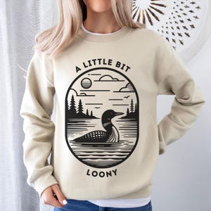 May include: A beige sweatshirt with a black graphic design of a loon in a lake with trees and the text "A Little Bit Loony".
