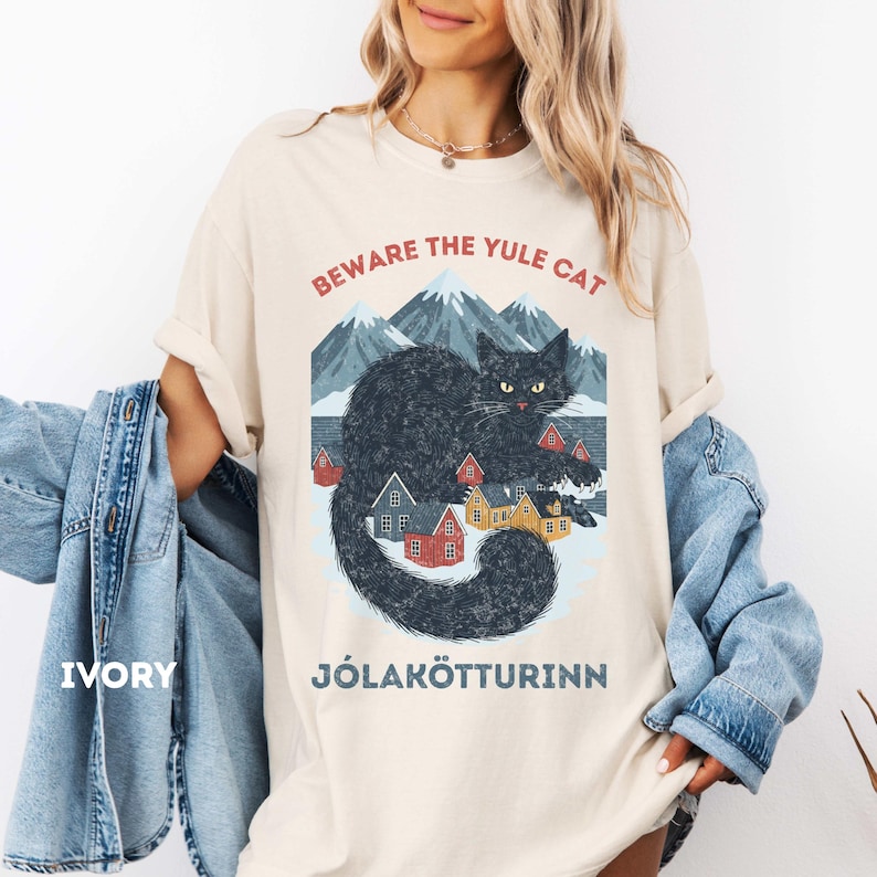 May include: Ivory-colored t-shirt with a graphic of a black cat, mountains, and small houses. Text reads "BEWARE THE YULE CAT" and "J&Oacute;LAK&Ouml;TTURINN". The shirt has a relaxed fit.