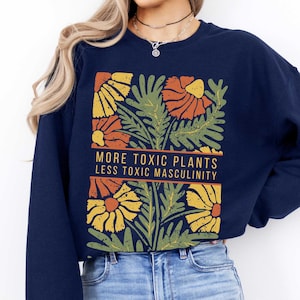 May include: A navy blue cropped sweatshirt with a graphic of stylized flowers and leaves in shades of orange, yellow, and green. The text "More Toxic Plants Less Toxic Masculinity" is printed in white on a black banner.