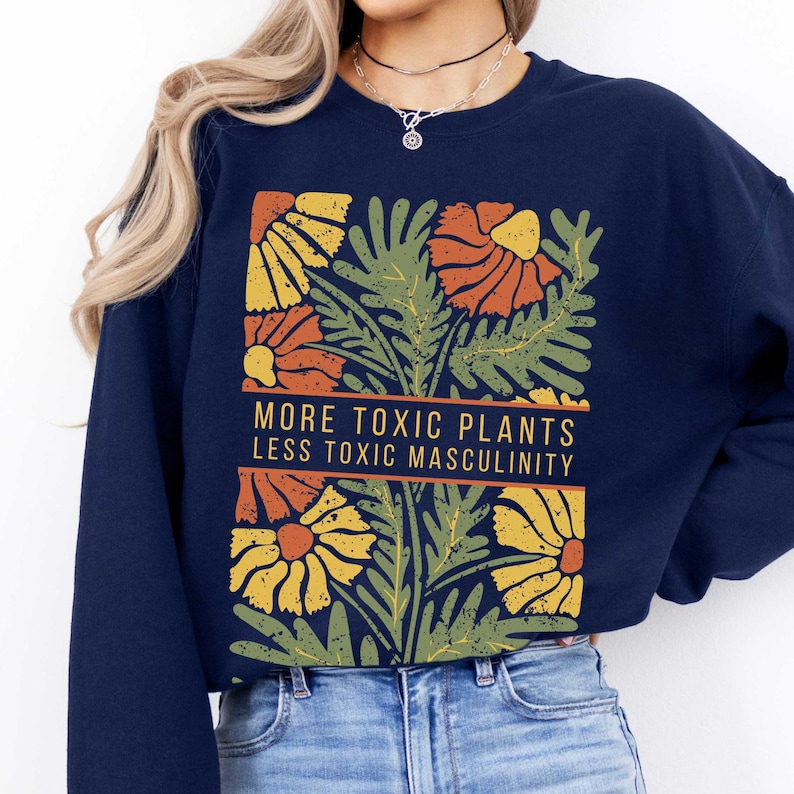 May include: A navy blue cropped sweatshirt with a graphic of stylized flowers and leaves in shades of orange, yellow, and green. The text "More Toxic Plants Less Toxic Masculinity" is printed in white on a black banner.