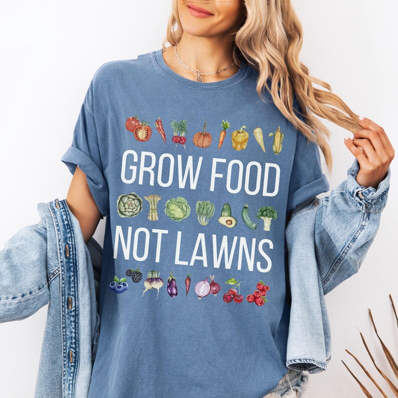 May include: A blue t-shirt with the text "GROW FOOD NOT LAWNS" in white. The shirt features watercolor illustrations of various vegetables and fruits, including tomatoes, carrots, and berries. The shirt is a comfortable, casual style.