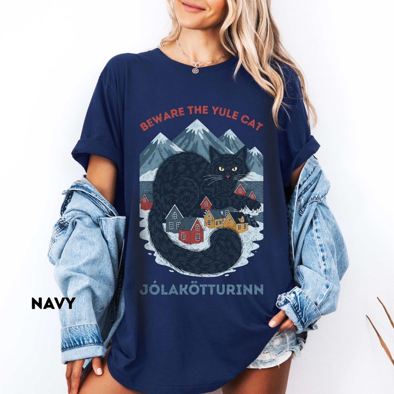 May include: Navy blue t-shirt featuring a graphic of a black cat with yellow eyes, mountains, and houses. The text "BEWARE THE YULE CAT" and "J&Oacute;LAK&Ouml;TTURINN" are printed above and below the cat illustration. The shirt is a relaxed fit.