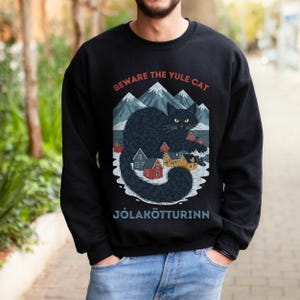 May include: Black sweatshirt with a graphic of a black cat, mountains, and colorful houses. The text "BEWARE THE YULE CAT" and "J&Oacute;LAK&Ouml;TTURINN" are printed above and below the graphic. The sweatshirt is a crew neck style.