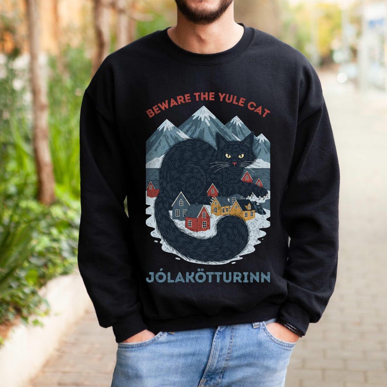 May include: Black sweatshirt with a graphic of a black cat, mountains, and colorful houses. The text "BEWARE THE YULE CAT" and "J&Oacute;LAK&Ouml;TTURINN" are printed above and below the graphic. The sweatshirt is a crew neck style.