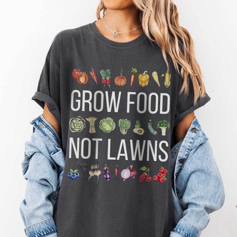May include: Dark gray t-shirt with the text "GROW FOOD NOT LAWNS" in white. The shirt features watercolor illustrations of various vegetables and fruits, including tomatoes, carrots, and berries, promoting gardening.