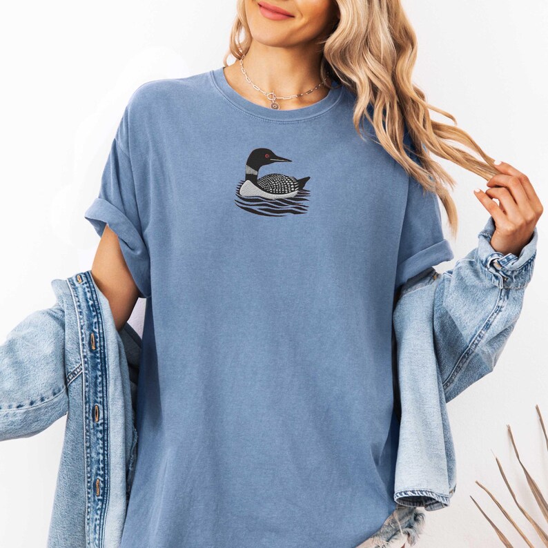 May include: A blue t-shirt with a black and white loon graphic. The loon is swimming in water with ripples.