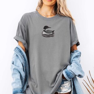 May include: A gray oversized t-shirt with a black and white loon graphic on the front. The loon is swimming in water with a wave pattern.