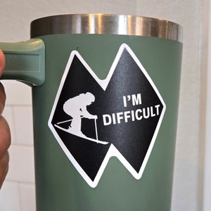 May include: A green stainless steel travel mug with a black and white sticker that says "I'm Difficult" and features a skier in a diamond shape.