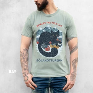 May include: Sage green t-shirt featuring a graphic of a black cat with yellow eyes, mountains, and small houses. The text "BEWARE THE YULE CAT" and "J&Oacute;LAK&Ouml;TTURINN" are printed above and below the cat illustration.