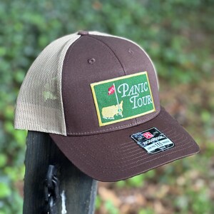 Peut inclure: Casquette de camionneur marron et beige avec un écusson "Panic Tour" orné d'un corgi et d'un drapeau de golf. La casquette a une visière incurvée et un dos en filet. L'étiquette de la marque Richardson est visible.