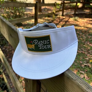 May include: White visor with the text "PANIC TOUR" on a green and gold patch. The visor has a curved brim and is made of a smooth, solid material. The visor is resting on a wooden fence.