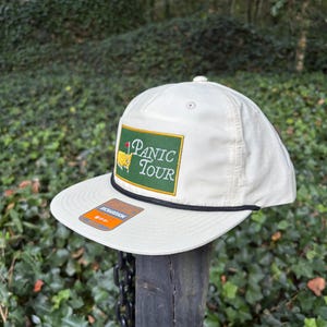 May include: Off-white baseball cap with a flat peak and a black rope detail. The cap has a green and gold patch that reads "PANIC TOUR" with a golf flag graphic. An orange tag is attached to the brim.