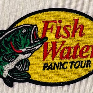 May include: An oval embroidered patch featuring a green and black fish on the left side. The background is yellow with a black border. The text "Fish Water" is stitched in red, and the text "PANIC TOUR" is stitched in black below it.