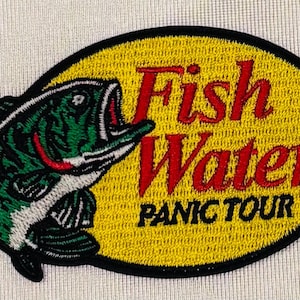 May include: An oval embroidered patch featuring a green and black fish on the left side. The background is yellow with a black border. The text "Fish Water" is stitched in red, and the text "PANIC TOUR" is stitched in black below it.