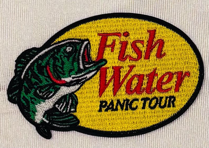 May include: An oval embroidered patch featuring a green and black fish on the left side. The background is yellow with a black border. The text "Fish Water" is stitched in red, and the text "PANIC TOUR" is stitched in black below it.