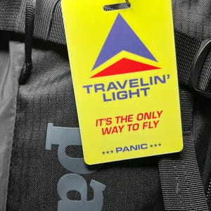 May include: A rectangular yellow luggage tag attached to a black backpack with a metal cable. The tag features a blue and red triangle logo, the text TRAVELIN' LIGHT, IT'S THE ONLY WAY TO FLY, and the word PANIC surrounded by three stars.