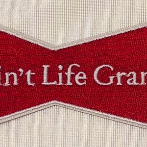 May include: A red, bow-tie shaped embroidered patch with a beige border. The text Ain't Life Grand is stitched in white across the center of the red fabric. The patch has a textured surface and a distinct, symmetrical, angular design.