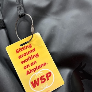 May include: A rectangular yellow luggage tag attached to a black bag with a metal wire loop. The tag features red text that reads Sitting around waiting on an Airplane. Below the text is a white circle containing the red letters WSP and 8-1962.
