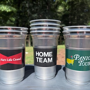 May include: Three silver-coloured plastic cups stacked together. The left cup features a black label with a red bowtie graphic and the text Ain't Life Grand. The centre cup has a black label with the text HOME TEAM. The right cup has a green label with a yellow map graphic and the text PANIC TOUR.
