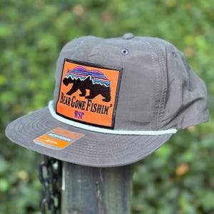 May include: Grey baseball cap with a flat peak and a white rope detail. The cap has a rectangular patch with a black bear silhouette, the text "Bear Gone Fishin'", and colourful accents on an orange background.