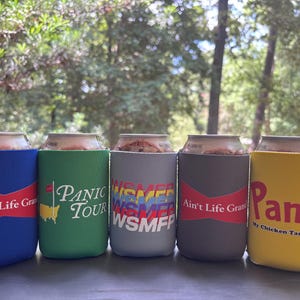 May include: Five cylindrical foam beverage insulators in blue, green, grey, dark grey, and yellow. The blue and dark grey ones feature a red bow-tie graphic with the text Ain't Life Grand. The green one shows a map and Panic Tour. The grey one displays WSMFP, and the yellow one features the word Panic with the text My Chicken Tastes Good.