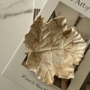 May include: A leaf-shaped decorative dish with a marbled beige and cream finish sits on a book titled "The Art of". The book's cover is white with black text, including the author's name, Shea McGee.