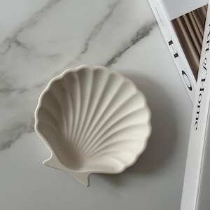 May include: A white, shell-shaped decorative dish. The dish has a ribbed texture and a small, pointed edge. The dish is made of ceramic or a similar material. The background is a marble-patterned surface.
