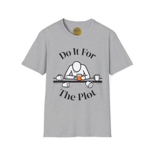 Puede incluir: Camiseta gris jaspeado con la frase "Do It For The Plot" en negro. El diseño muestra una figura de palo en una mesa con tazas de café. La camiseta tiene cuello redondo y mangas cortas.
