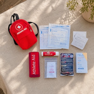 May include: A red Athlete Aid mini backpack containing medical supplies including two McKesson non-woven sterile sponges, alcohol prep pads, a red Athlete Aid lip balm, a packet of triple antibiotic ointment, an Athlete Aid card, and a Mela sunscreen stick.