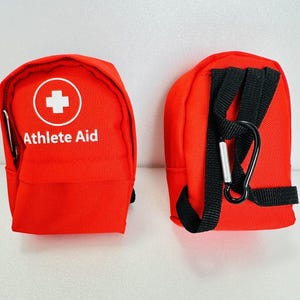 May include: Two bright red fabric pouches with black trim and black webbing straps. The front of one pouch features a white circular logo with a cross and the text Athlete Aid. The other pouch shows black straps and a black metal carabiner clip.