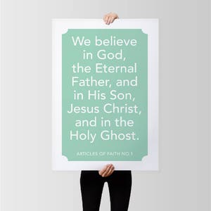 May include: A rectangular white framed poster with a mint green background and white text. The text reads "We believe in God, the Eternal Father, and in His Son, Jesus Christ, and in the Holy Ghost." The text "ARTICLES OF FAITH NO. 1" is at the bottom.