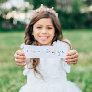 May include: A young girl wearing a crown holds a white card with the words "i am a child of god" written in rainbow colors.