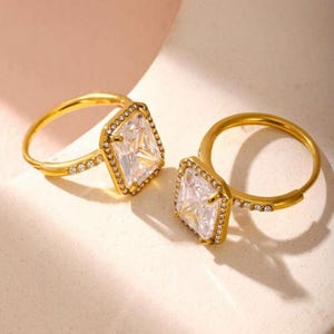 Puede incluir: Dos anillos dorados con grandes piedras preciosas cuadradas y transparentes. Las piedras preciosas están rodeadas de piedras más pequeñas y transparentes. Las bandas de los anillos también están adornadas con pequeñas piedras transparentes.