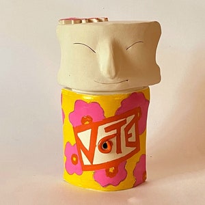 May include: A ceramic figurine with a smiling face and a yellow body with pink flowers and the word "Vote" in red. The figurine has a small hole in the bottom.