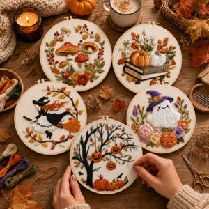 Op de afbeelding: Vijf ronde borduurringen met herfstthema's, waaronder paddenstoelen, pompoenen op boeken, een heks op een bezemsteel, een geest met een hoed en een boom met hangende pompoenlantaarns, omringd door losse draad en herfstbladeren.