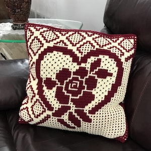 May include: A square crochet cushion featuring a cream-coloured background and a burgundy heart-shaped floral design. The cushion is on a brown leather sofa. The cushion's border is burgundy.