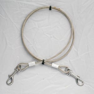 May include: A silver-colored dog tie-out cable with two metal swivel clips. The cable is made of braided steel and is secured with black plastic fasteners. The clips are designed to attach to a dog's collar or harness.