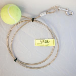 May include: A dog toy featuring a bright yellow tennis ball attached to a silver-colored cable leash. The leash has a metal clasp and a small yellow tag that reads "ball-stop". The cable is looped and secured with black fasteners.